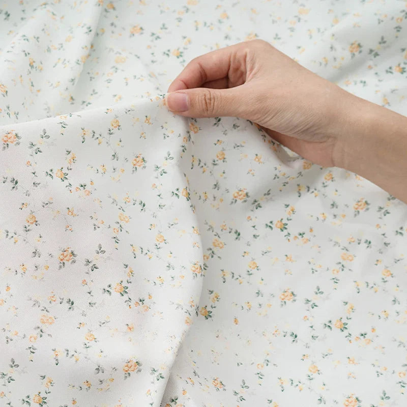 Une main tient le tissu mousseline à fleurs au mètre de Longan Craft France, un tissu imprimé en polyester léger avec un petit motif floral jaune et vert, doucement drapé sur une surface plane - idéal pour la création de vêtements.