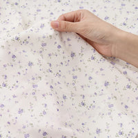 Une main tient un morceau de tissu mousseline à fleurs au mètre de Longan Craft France, un tissu imprimé en polyester doux avec de délicats motifs floraux violet clair et vert, parfait pour créer des vêtements élégants.