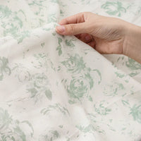 A hand lifts a piece of tissu mousseline à fleurs au mètre de Longan Craft France, dévoilant la texture délicate et le motif floral vert clair sur ce tissu mousseline polyester imprimé idéal pour la confection de vêtements.