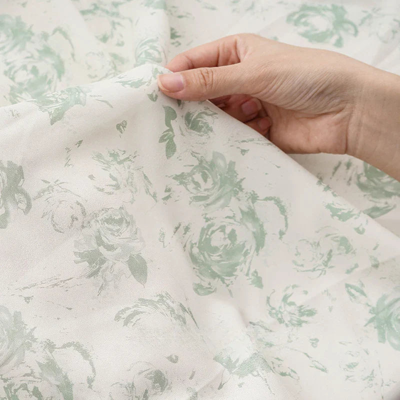 A hand lifts a piece of tissu mousseline à fleurs au mètre de Longan Craft France, dévoilant la texture délicate et le motif floral vert clair sur ce tissu mousseline polyester imprimé idéal pour la confection de vêtements.