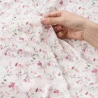 Une main tient délicatement le tissu mousseline à fleurs au mètre de Longan Craft France, révélant son polyester doux et aéré et son délicat motif floral rose et gris - un tissu imprimé parfait pour la création de vêtements.