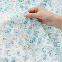 Une main tient et soulève le Tissu mousseline à fleurs au mètre de Longan Craft France, dévoilant la délicatesse du tissu polyester imprimé aux motifs floraux bleu et vert, idéal pour la confection de vêtements.