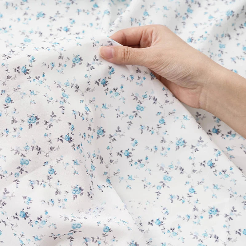 Une main tient et soulève un morceau de Tissu mousseline à fleurs au mètre de Longan Craft France, un tissu léger en polyester aux délicats imprimés floraux bleus et gris, idéal pour l'habillement et la couture.