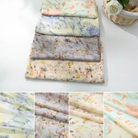 Cinq pièces de Tissu mousseline à fleurs au mètre de Longan Craft France sont empilées sur une surface claire près d'un panier de fleurs blanches. Les gros plans montrent les délicats motifs pastel et la texture de cette mousseline en polyester pour confection de vêtements.