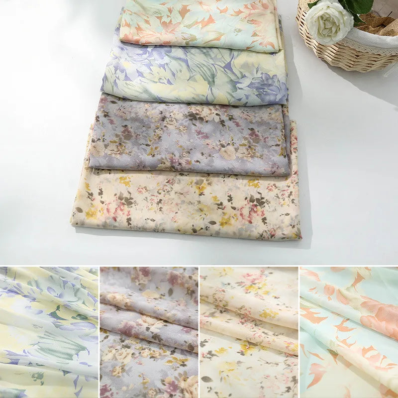 Cinq pièces de Tissu mousseline à fleurs au mètre de Longan Craft France sont empilées sur une surface claire près d'un panier de fleurs blanches. Les gros plans montrent les délicats motifs pastel et la texture de cette mousseline en polyester pour confection de vêtements.