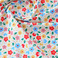 Un gros plan de Popeline de coton à fleurs au mètre de Longan Craft France montre son imprimé floral doux 100% coton avec des fleurs rouges, bleues, jaunes et des feuilles vertes sur fond blanc - tissu idéal pour le matelassage, les langes, les peignoirs et les chemises.
