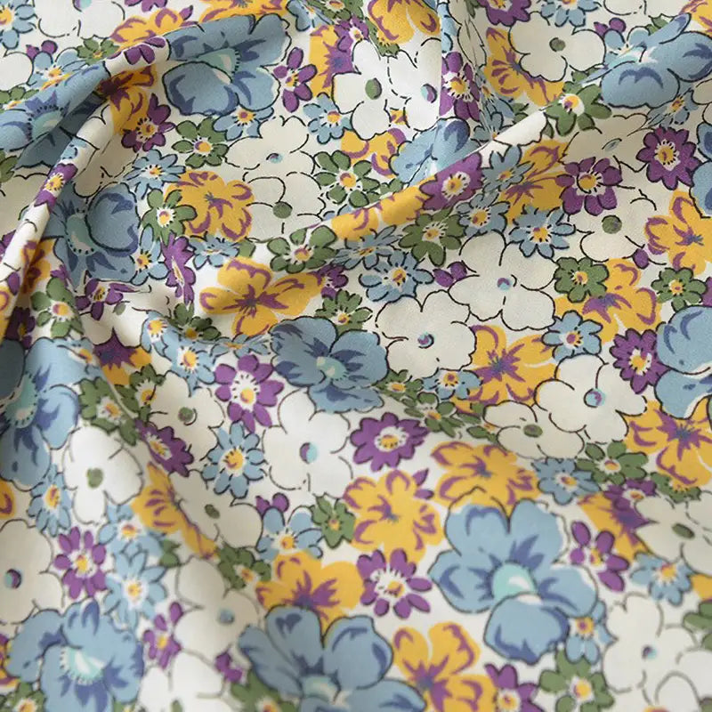 Un gros plan de la "Popeline de coton à fleurs au mètre" de Longan Craft France montre une popeline florale douce et légèrement froissée avec des fleurs bleues, jaunes, violettes et blanches sur un fond clair - idéale pour le quilting, les nappes, les robes et les chemises.