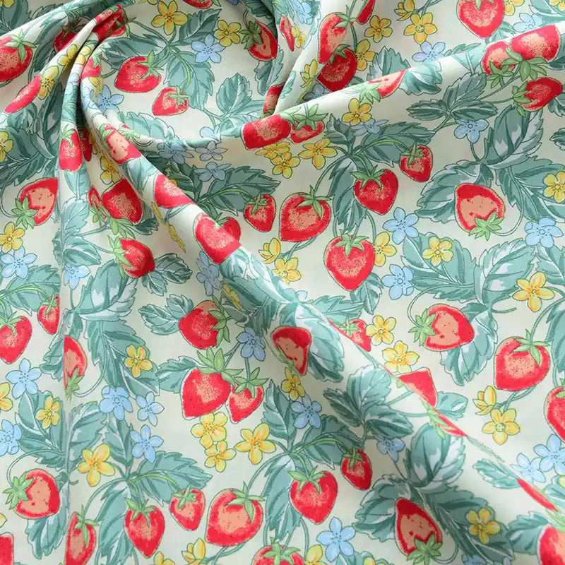 Un gros plan de Popeline de coton à fleurs au mètre de Longan Craft France montre son tissu doux 100% coton, avec des fraises rouges, des feuilles vertes et des fleurs bleues/jaunes sur un fond clair - idéal pour le quilting, les langes, les peignoirs et les chemises.