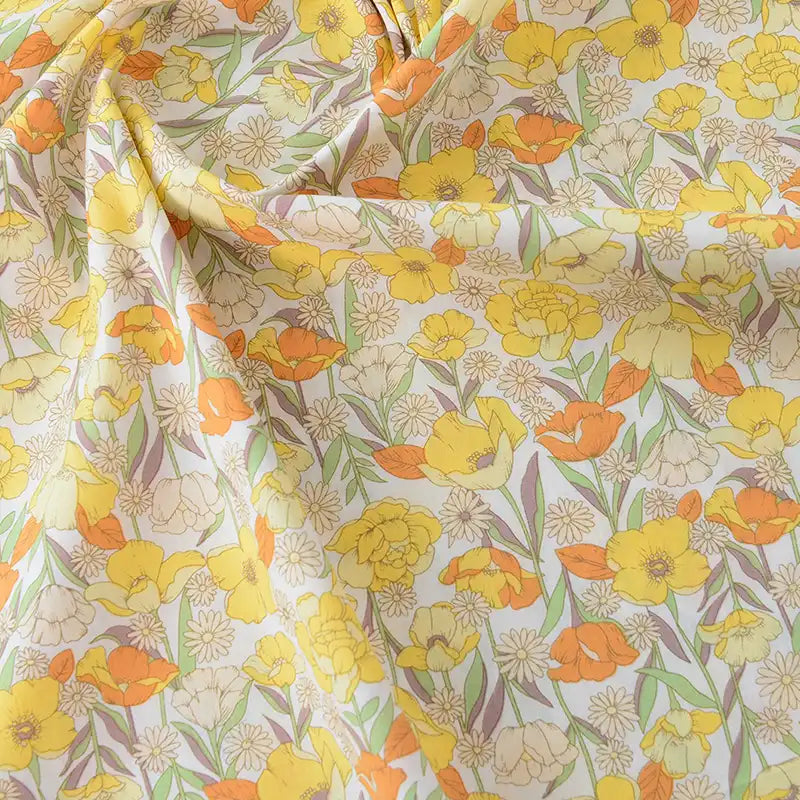 Gros plan sur la Popeline de coton à fleurs au mètre de Longan Craft France, montrant des fleurs jaunes et orange avec des feuilles vertes sur un fond crème. Ce tissu 100% coton, doux et légèrement brillant, est idéal pour le quilting, les nappes, les robes et les chemises.