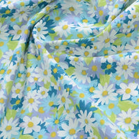 Gros plan sur la Popeline de coton à fleurs au mètre de Longan Craft France, un tissu floral 100% coton avec des marguerites blanches et jaunes, des feuilles vertes et des fleurs bleues sur fond bleu clair - idéal pour le quilting, les langes, les peignoirs et les chemises.