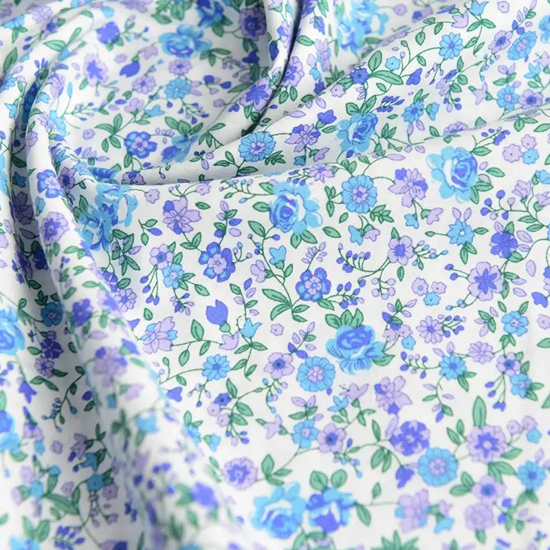 Un gros plan de Popeline de coton à fleurs de Longan Craft France montre un tissu floral doux, 100 % coton, avec des fleurs bleues et violettes et des feuilles vertes, légèrement drapé - idéal pour le quilting, les nappes, les robes ou les chemises.