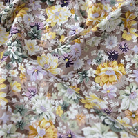 Gros plan sur la Popeline de coton à fleurs de Longan Craft France, qui présente des motifs floraux jaunes, blancs et violets avec des feuilles vertes sur un fond marron clair. Ce doux tissu 100% coton est parfait pour le quilting, les langes, les peignoirs et les chemises.
