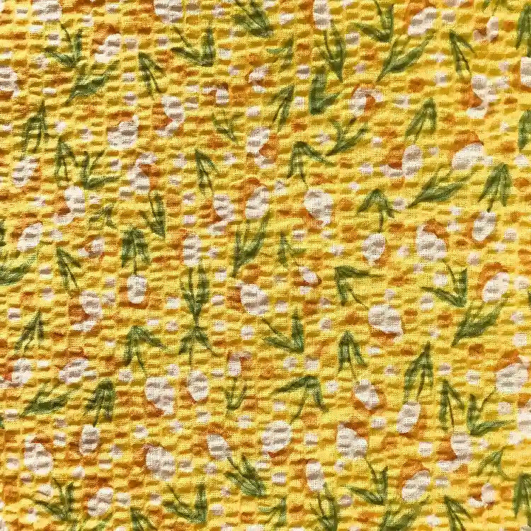 Le Tissu seersucker fleuri au mètre de Longan Craft France est un seersucker de coton vintage jaune avec de petites feuilles vertes et des grappes de points blancs dans un motif floral répétitif et uniformément réparti.