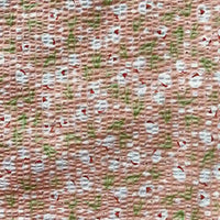 Un gros plan du tissu seersucker fleuri au mètre de Longan Craft France montre son coton rose, léger et texturé avec de petites fleurs blanches, des tiges vertes et des accents rouges - parfait comme tissu imprimé froissé d'inspiration vintage.
