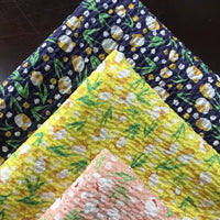 Quatre pièces de Tissu seersucker fleuri au mètre de Longan Craft France, un coton imprimé de style vintage de la collection Tissu seersucker, sont empilées en bleu foncé, jaune vif, jaune clair et rose pâle sur un fond sombre.