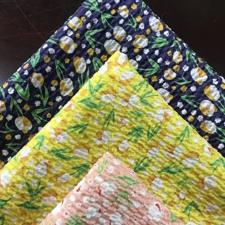 Quatre pièces de Tissu seersucker fleuri au mètre de Longan Craft France, un coton imprimé de style vintage de la collection Tissu seersucker, sont empilées en bleu foncé, jaune vif, jaune clair et rose pâle sur un fond sombre.