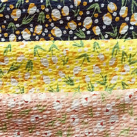 Quatre bandes texturées du tissu seersucker fleuri au mètre de Longan Craft France présentent des imprimés floraux et ovoïdes en jaune, blanc et vert sur du coton bleu, jaune, jaune clair et pêche à effet vintage pour un look léger et élégant.