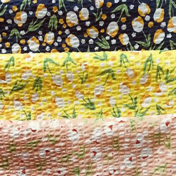Quatre bandes texturées du tissu seersucker fleuri au mètre de Longan Craft France présentent des imprimés floraux et ovoïdes en jaune, blanc et vert sur du coton bleu, jaune, jaune clair et pêche à effet vintage pour un look léger et élégant.
