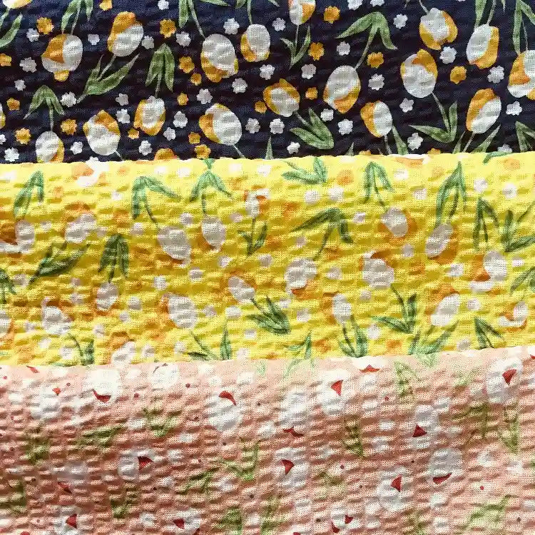 Quatre bandes texturées du tissu seersucker fleuri au mètre de Longan Craft France présentent des imprimés floraux et ovoïdes en jaune, blanc et vert sur du coton bleu, jaune, jaune clair et pêche à effet vintage pour un look léger et élégant.