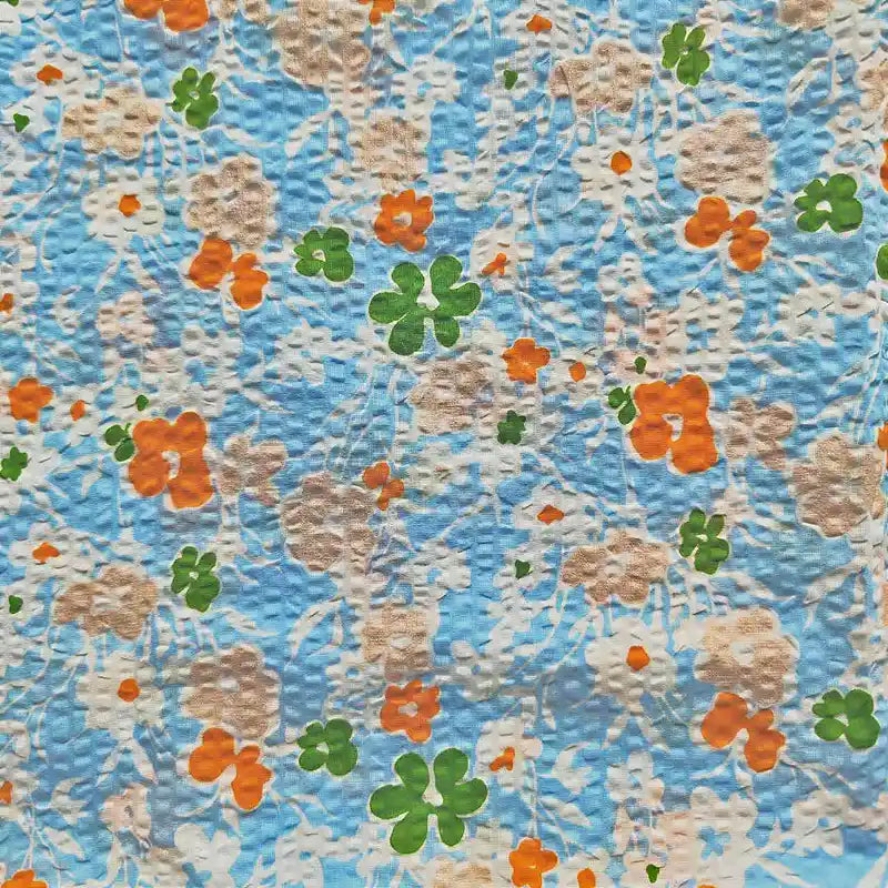 Le Tissu seersucker imprimé floral au mètre de Longan Craft France est un coton seersucker bleu texturé avec un motif floral vintage gaufré en orange, beige et vert.