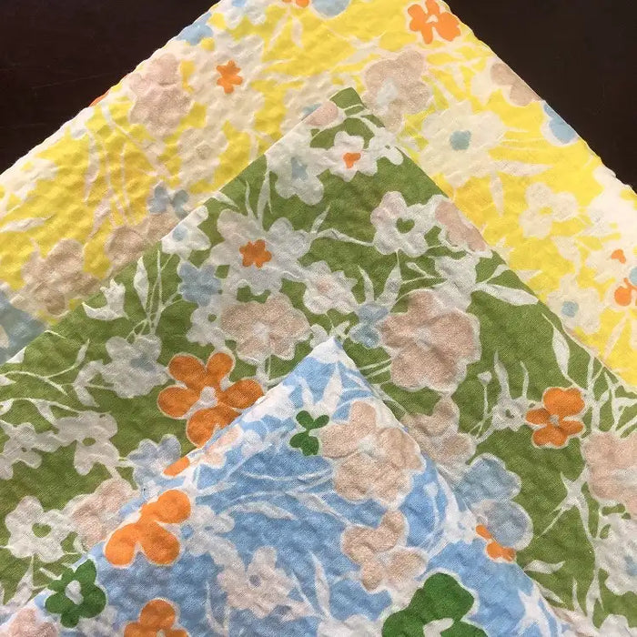 Trois pièces de Tissu seersucker imprimé floral au mètre de Longan Craft France en jaune, vert et bleu avec des fleurs multicolores se superposent sur un fond marron foncé, mettant en valeur un tissu de coton léger et texturé de style vintage.