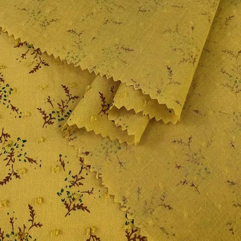 Plumetis en voile jacquard jaune de Longan Craft France, à la texture subtilement pointillée et à l'imprimé floral de fines branches feuillues. Bords coupés en zigzag, parfaits pour les robes d'été. Tissu au mètre pour confection et couture.