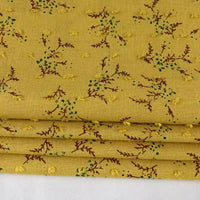 Un gros plan du Tissu voile jacquard plumetis floral au mètre de Longan Craft France montre un tissu jaune avec de petites branches brunes, des points verts et des points jaunes brodés, parfait pour les projets de couture et de confection.