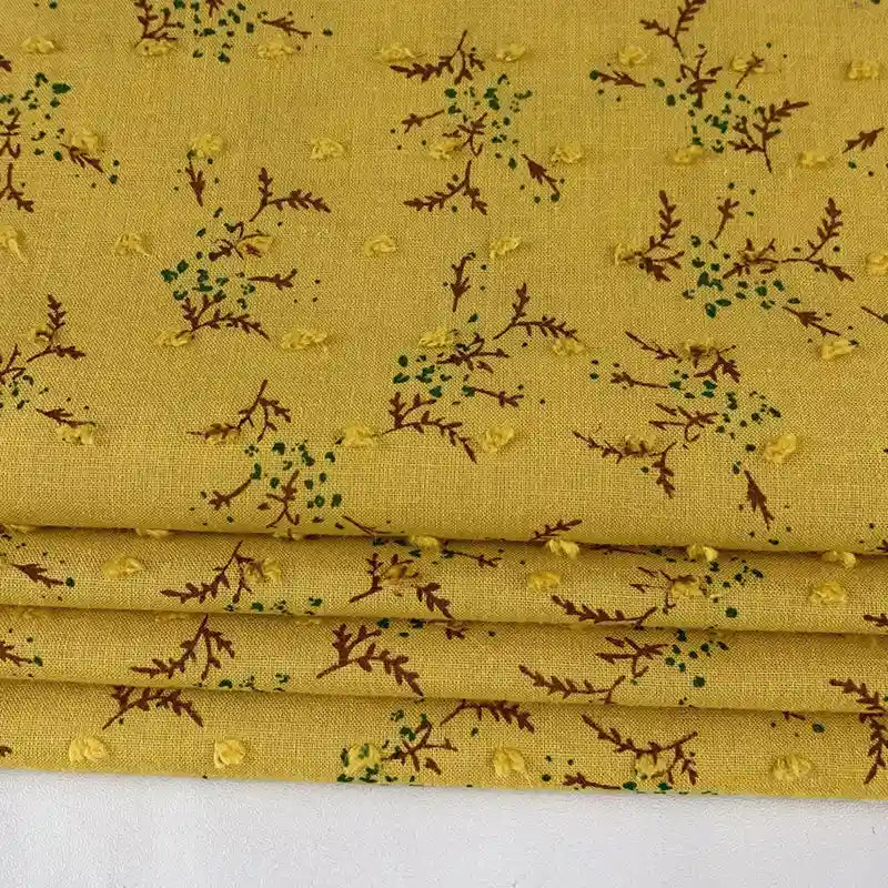 Un gros plan du Tissu voile jacquard plumetis floral au mètre de Longan Craft France montre un tissu jaune avec de petites branches brunes, des points verts et des points jaunes brodés, parfait pour les projets de couture et de confection.