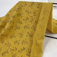 Un morceau de Tissu voile jacquard plumetis floral au mètre de Longan Craft France, comprenant un voile de coton jaune avec des motifs floraux bruns et verts, est drapé sur une surface blanche. Léger, avec des bords coupés en zigzag, il est idéal pour la confection de vêtements d'été.