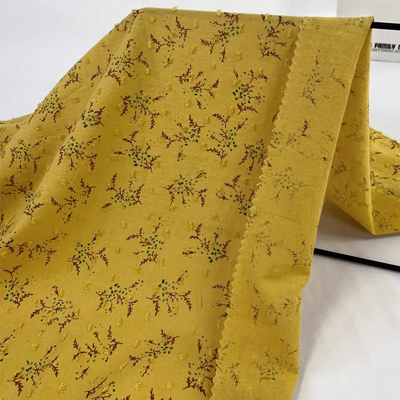 Un morceau de Tissu voile jacquard plumetis floral au mètre de Longan Craft France, comprenant un voile de coton jaune avec des motifs floraux bruns et verts, est drapé sur une surface blanche. Léger, avec des bords coupés en zigzag, il est idéal pour la confection de vêtements d'été.