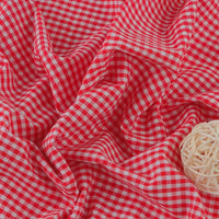 Le Tissu popeline vichy au mètre de Longan Craft France, un tissu léger à carreaux 100 % coton, est représenté froissé sur une surface avec une boule décorative en rotin crème placée dans le coin inférieur droit.