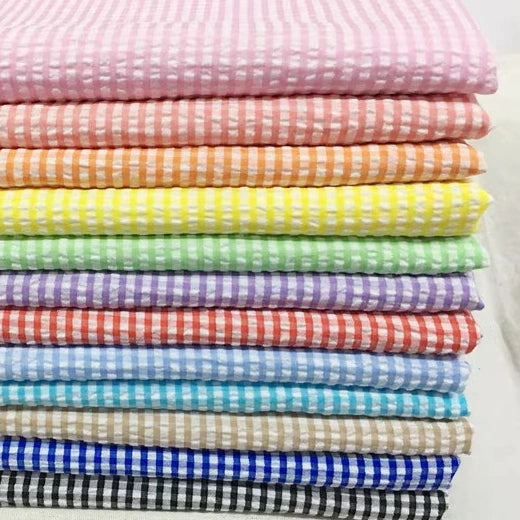 Une pile de tissu seersucker vichy de Longan Craft France, avec des carreaux classiques dans les tons rose, orange, jaune, vert, violet, bleu, rouge, beige, noir et blanc. Vendu au mètre en tant que tissu seersucker froncé.