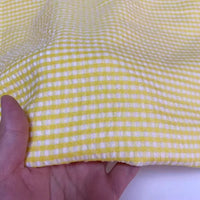 Une main présente le seersucker vichy au mètre de Longan Craft France, un tissu léger et froissé à carreaux jaunes et blancs, idéal pour une tenue décontractée ou estivale.