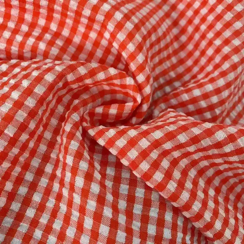 Le Seersucker vichy au mètre de Longan Craft France présente des carreaux rouges et blancs (motif à carreaux) ; le tissu seersucker froissé crée des plis et des ombres visibles, mettant en valeur sa texture légère et froncée.
