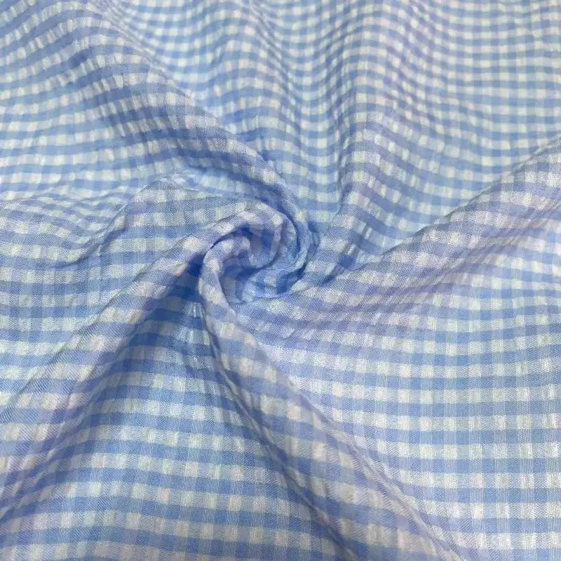 Un gros plan du Seersucker vichy au mètre de Longan Craft France montre un tissu seersucker bleu clair et blanc avec un motif à carreaux, tourbillonné au centre pour un aspect texturé et froissé.