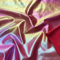Lamé irisé au mètre de Longan Craft France : tissu polyester lamé métallique brillant, effet chatoyant, reflets roses, violets, jaunes et dorés, partiellement drapé et plissé avec un coin rouge visible. Idéal pour costumes de scène et à coudre.