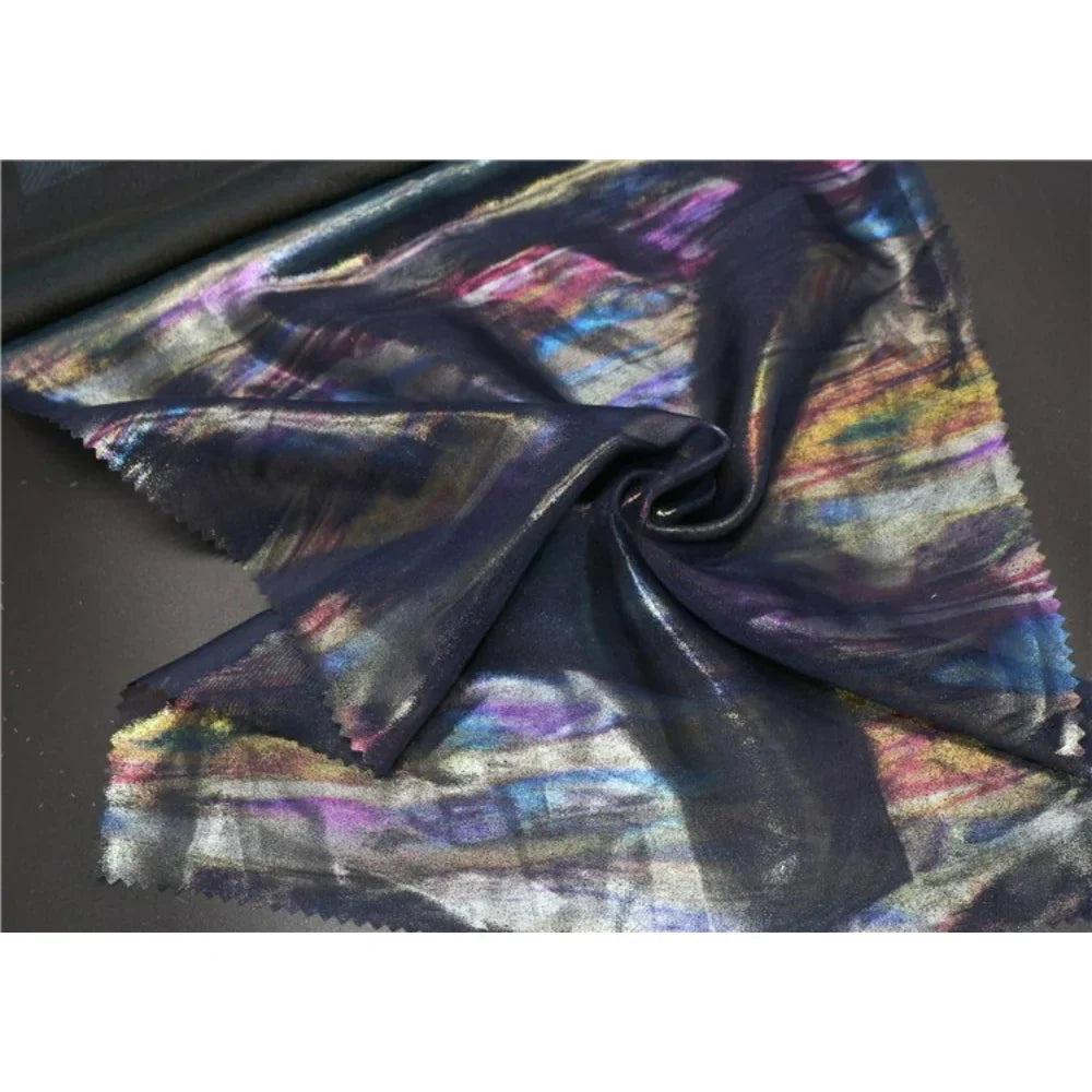 Un morceau de Mousseline irisée au mètre de Longan Craft France, une mousseline de polyester foncé métallisée avec un motif multicolore irisé, est étalé sur une table avec une partie torsadée en une spirale lâche au centre.