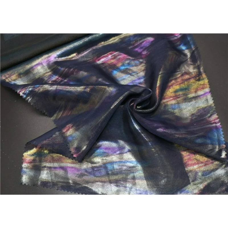 Un morceau de Mousseline irisée au mètre de Longan Craft France, une mousseline de polyester foncé métallisée avec un motif multicolore irisé, est étalé sur une table avec une partie torsadée en une spirale lâche au centre.