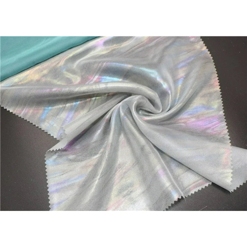 Un échantillon de Mousseline irisée au mètre de Longan Craft France est présenté en polyester métallisé gris clair avec une brillance arc-en-ciel subtile, des bords découpés au laser, un pli central et une bande de turquoise en haut à gauche.
