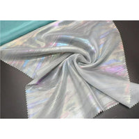 Un échantillon de Mousseline irisée au mètre de Longan Craft France est présenté en polyester métallisé gris clair avec une brillance arc-en-ciel subtile, des bords découpés au laser, un pli central et une bande de turquoise en haut à gauche.