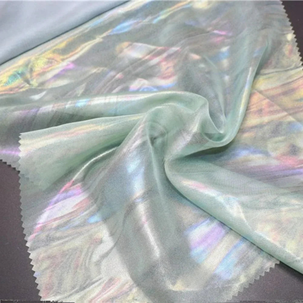 Un morceau de Mousseline irisée au mètre de Longan Craft France est disposé sur une surface sombre, rassemblé au centre pour mettre en valeur sa texture polyester brillante et semi-transparente et ses douces teintes pastel, parfaites pour les projets de bricolage.