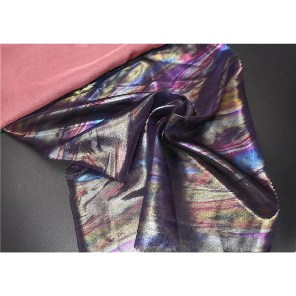 Un morceau de Mousseline irisée au mètre de Longan Craft France, une mousseline de polyester multicolore et brillante, est drapé sur un tissu noir à côté d'un tissu rose uni, reflétant des nuances de bleu, de violet et d'or - parfait pour les fournitures de bricolage.