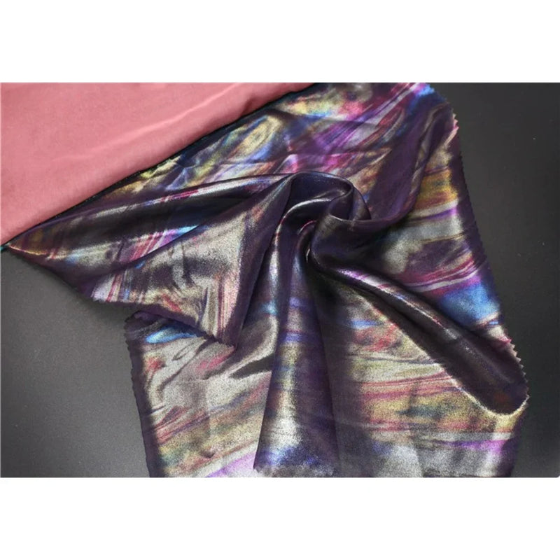 Un morceau de Mousseline irisée au mètre de Longan Craft France, une mousseline de polyester multicolore et brillante, est drapé sur un tissu noir à côté d'un tissu rose uni, reflétant des nuances de bleu, de violet et d'or - parfait pour les fournitures de bricolage.