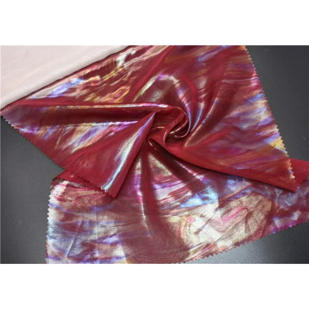 Un morceau de Mousseline irisée au mètre de Longan Craft France, une mousseline de polyester rouge brillant aux reflets multicolores tourbillonnants, est étalé sur une surface sombre, le bord supérieur replié. Parfaite pour le bricolage et l'artisanat.
