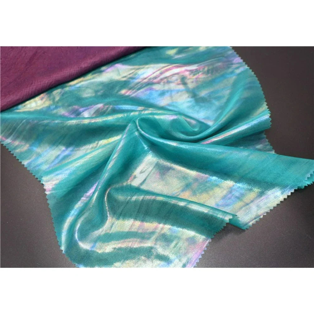 Une section de Mousseline irisée au mètre de Longan Craft France, une mousseline métallisée brillante en polyester sarcelle, est partiellement pliée à côté d'un tissu violet sur un fond sombre - parfait pour les projets de bricolage.