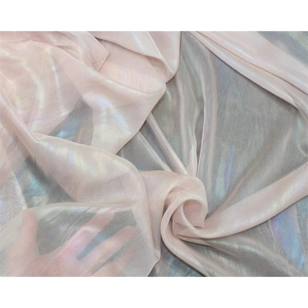 Un gros plan de la Mousseline irisée au mètre de Longan Craft France en rose pâle montre un tissu de polyester translucide et métallique doucement plié avec une lumière réfléchie et une main visible sous le textile transparent.