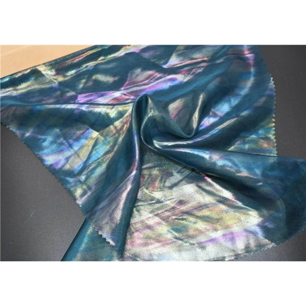 Un morceau de Mousseline irisée au mètre de Longan Craft France, une mousseline métallique en polyester brillant dans des tons bleus, verts et violets, est étalé sur une surface sombre avec une texture réfléchissante et des plis ondulés.