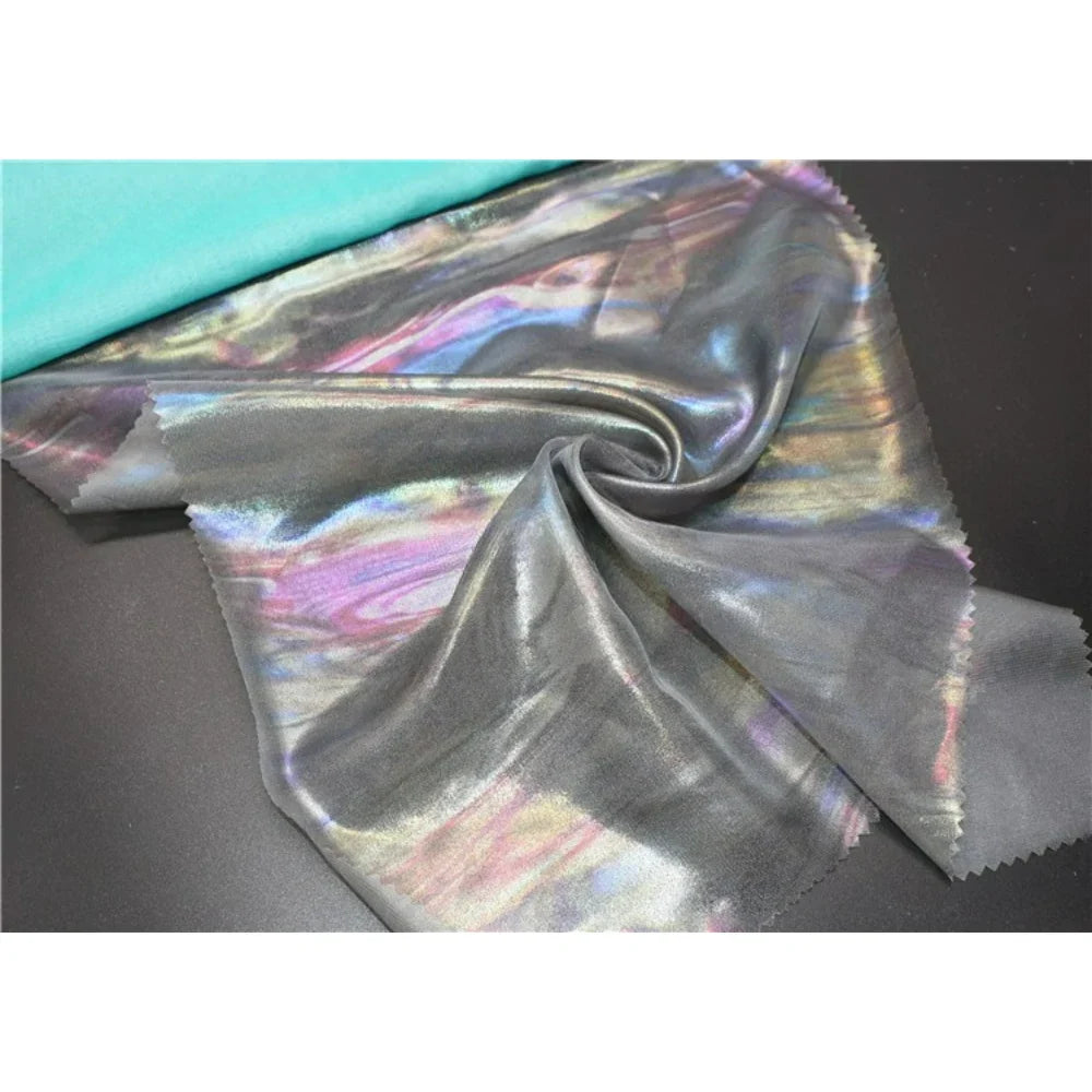 Un morceau de Mousseline irisée au mètre (tissu mousseline polyester métallisé) de Longan Craft France est étalé et légèrement froncé au centre, montrant un tissu turquoise uni sur le bord supérieur sur un fond noir.