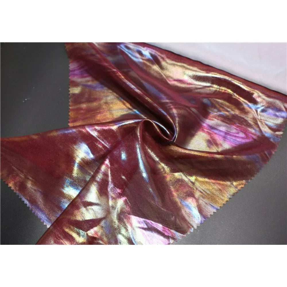Un morceau de Mousseline irisée au mètre de Longan Craft France, un tissu de polyester rouge et violet chatoyant, est étalé sur une surface sombre. Ses reflets métalliques reflètent la lumière en différentes couleurs et les bords coupés en zigzag apportent une touche raffinée.