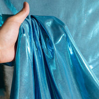 Une main tient le Lamé métallique liquide au mètre de Longan Craft France sur un fond bleu. Ce tissu lisse, brillant et métallique est parfait pour les costumes de scène, les robes de soirée et la couture.